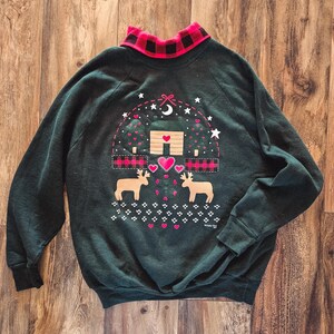 May include: Dark green sweatshirt with a red and black plaid collar. The front features a cabin scene with deer, hearts, and stars. The cabin has a heart-shaped door and is surrounded by trees.