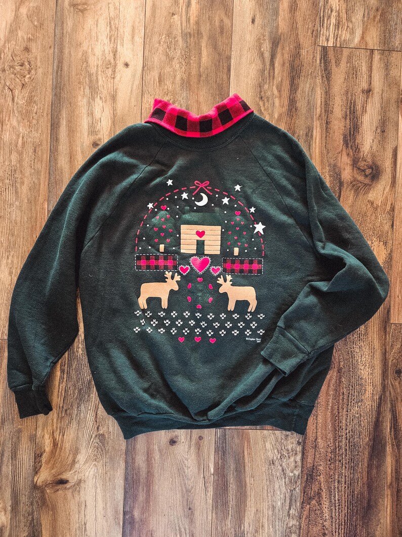 May include: Dark green sweatshirt with a red and black plaid collar. The front features a cabin scene with deer, hearts, and stars. The cabin has a heart-shaped door and is surrounded by trees.