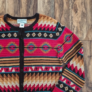 Peut inclure: Un cardigan à boutons avec un motif géométrique coloré. Le pull présente un col et une patte de boutonnage noirs, avec un motif dans les tons de rouge, noir, beige et turquoise. La marque "Pendleton" est visible.