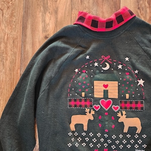 May include: Dark green sweatshirt with a red and black plaid collar. The front features a cabin, trees, hearts, stars, and two reindeer. The design is outlined with a dotted line. The sweatshirt has long sleeves.