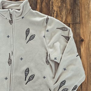 May include: A light gray fleece jacket with a full zipper and a high collar. The jacket is patterned with black snowshoe and snowflake designs. The jacket is made of a soft, warm material.