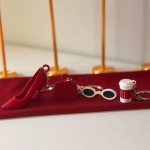 Puede incluir: Colección de llaveros rojos, que incluyen un zapato de tacón alto, un bolso, gafas de sol y una taza de café. Los artículos están colocados sobre una alfombra roja, con postes dorados en el fondo.