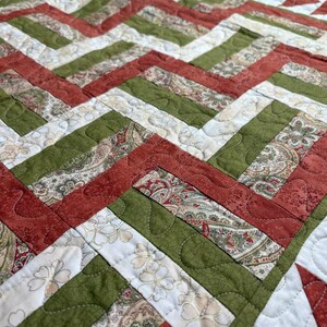 May include: A handmade quilt with a geometric chevron pattern. The quilt features alternating bands of red, green, and white fabric. The white fabric has a floral pattern, while the red and green fabrics have a paisley design. The quilt is machine-stitched.