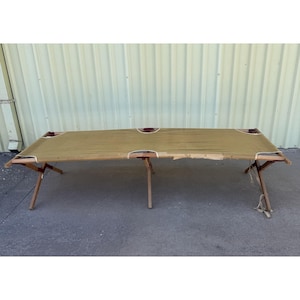 May include: A vintage folding cot with a khaki canvas bed and wooden frame. The cot has a rectangular shape with rounded corners and is supported by folding legs. The canvas shows signs of wear, adding to its rustic charm. A classic piece for camping or guest use.