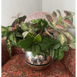 Puede incluir: Una maceta esférica de metal plateado pulido que contiene un denso arreglo de follaje artificial. La planta presenta una mezcla de hojas de color verde sólido y hojas variegadas con tonos rosados, crema y verde oscuro en los tallos.