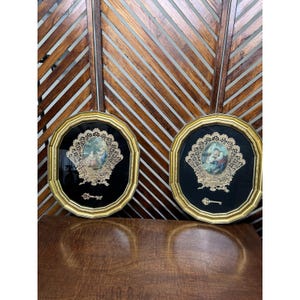 May include: Two framed oval artworks with gold trim. Each features a central image of a religious scene, surrounded by ornate gold lace on a black background. A small gold key is below the image. The frames are displayed against a wooden slatted backdrop.