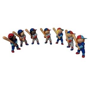 Puede incluir: Una colección de ocho figuras de jugadores de béisbol. Cada figura lleva un uniforme de béisbol con gorra y sostiene un bate de madera. Los uniformes son de varios colores, incluyendo rojo, azul y gris. Las figuras están dispuestas en semicírculo.
