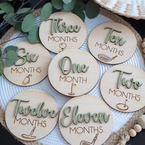 May include: Wooden monthly milestone discs with laser-etched golf-themed designs. Each disc features a number in a green font, indicating the month. The discs are arranged on a white surface with a woven texture, with eucalyptus leaves in the background.