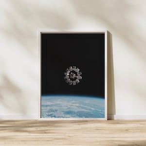 May include: Framed print featuring a detailed depiction of a space station in orbit above Earth. The artwork showcases a circular, metallic spacecraft against a backdrop of deep space and the planet's atmosphere. The print is displayed in a white frame.