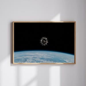 May include: A framed print depicting a space station in orbit above Earth. The space station is silver and circular, with multiple modules. The Earth below is blue and white, with visible cloud formations. The background is black.