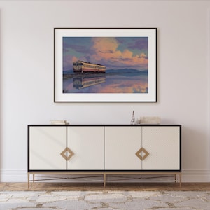 May include: Framed art print of a train on a body of water, reflecting the sky. The train is cream and red, set against a backdrop of a colorful sunset with pink and blue hues. The artwork is displayed above a white and black cabinet.