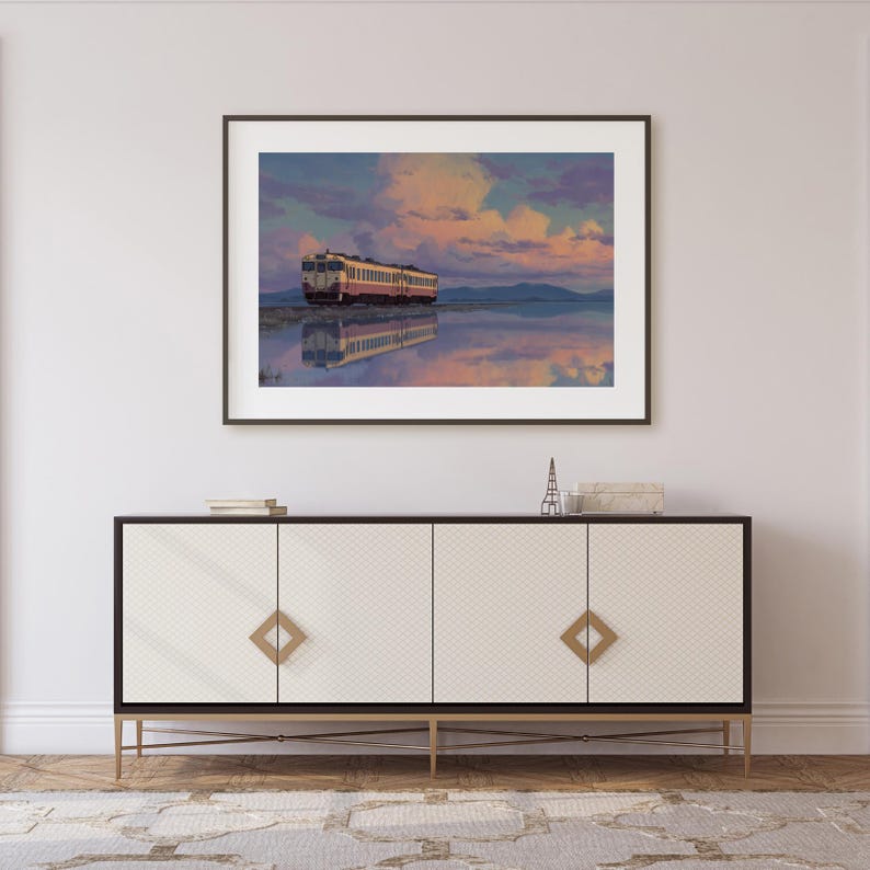 May include: Framed art print of a train on a body of water, reflecting the sky. The train is cream and red, set against a backdrop of a colorful sunset with pink and blue hues. The artwork is displayed above a white and black cabinet.