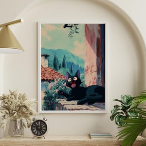 May include: A framed art print featuring a black cat with wide eyes, lounging on a stone ledge. The background depicts a scenic view of a European village with red-tiled roofs, mountains, and a blue sky. The artwork has a whimsical, animated style.