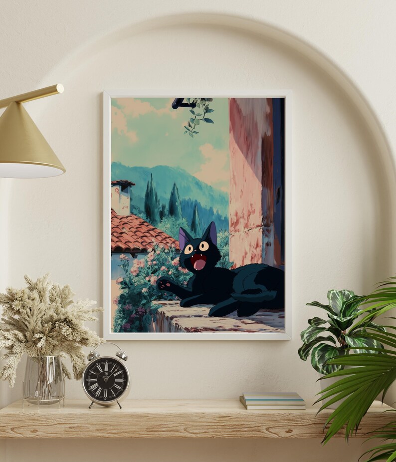 May include: A framed art print featuring a black cat with wide eyes, lounging on a stone ledge. The background depicts a scenic view of a European village with red-tiled roofs, mountains, and a blue sky. The artwork has a whimsical, animated style.