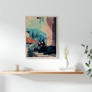 May include: A framed art print featuring a black cat with wide eyes, set against a backdrop of a European village scene. The cat is lying on a stone ledge, with a view of mountains and buildings. The artwork has a vintage, animated style.