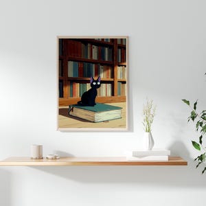 May include: A framed print featuring a black cat with large eyes sitting atop a teal book. The background shows a library with bookshelves filled with books. The print has a light wood frame and is displayed on a white wall.