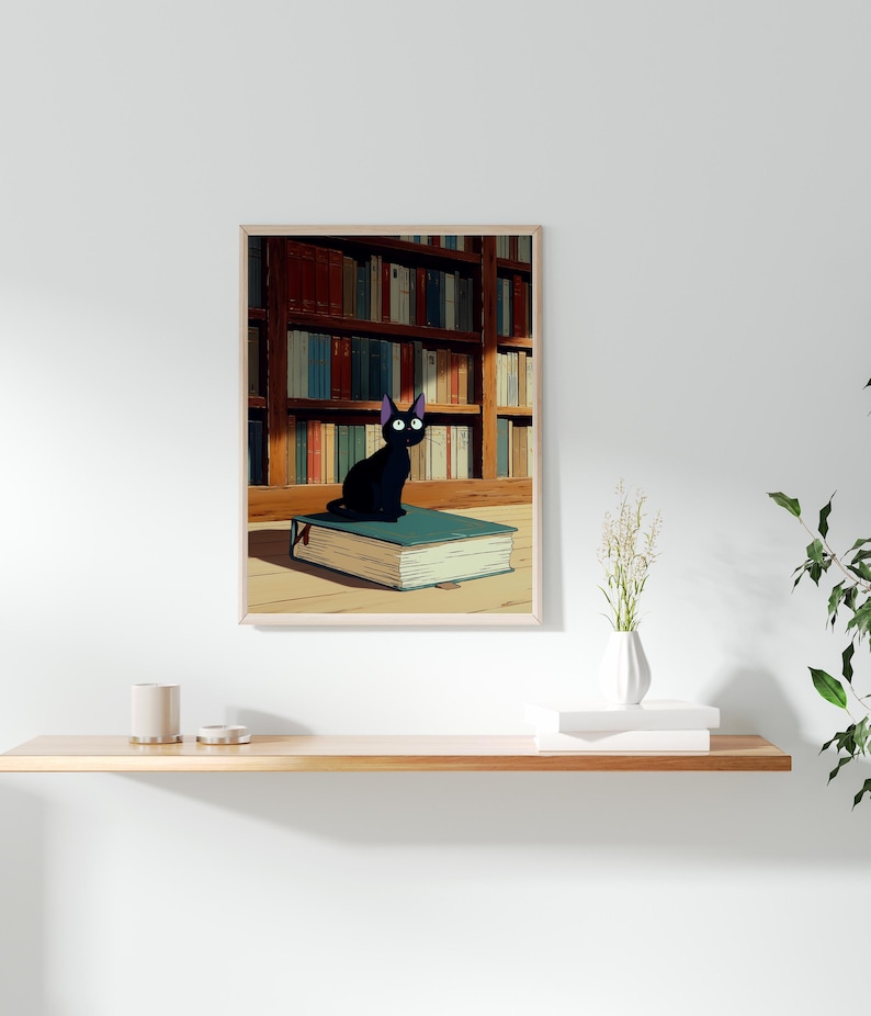 May include: A framed print featuring a black cat with large eyes sitting atop a teal book. The background shows a library with bookshelves filled with books. The print has a light wood frame and is displayed on a white wall.
