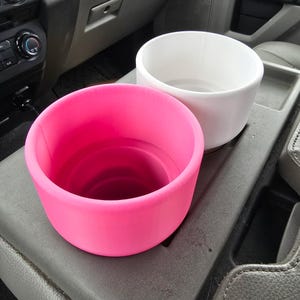 May include: Two car cup holders, one bright pink and one white, sit side-by-side in a vehicle's console. The cup holders are cylindrical with a smooth, matte finish. The interior of the pink cup holder is visible.