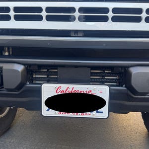May include: Close-up of the front of a white Ford Bronco. The word "FORD" is displayed in red letters across the top grille. A California license plate is mounted below the grille, with the word "California" visible.