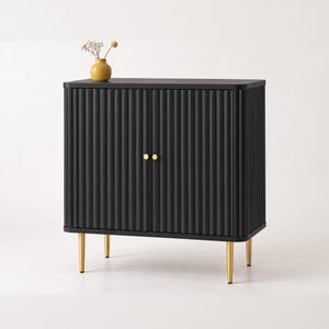 May include: A black cabinet with a fluted, ribbed texture on the two front doors. The cabinet features two small, round gold-toned knobs and is supported by four slender, tapered gold-toned metal legs. A small yellow vase sits on top.