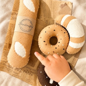 May include: A collection of felt food toys, including a baguette with a "Handcrafted" label, a bagel, a croissant, and a chocolate donut. The toys are arranged on a piece of paper with printed text.