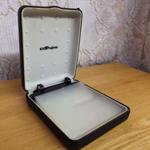 May include: A black rectangular storage case with a hinged lid, featuring a white interior with the text cdProjects printed in black. The case contains multiple clear plastic sleeves for organizing media discs, held in place by a black plastic mechanism.