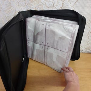 May include: A black zippered binder featuring multiple clear plastic sleeves with white paper inserts. The binder is open, revealing the internal storage pages designed for organizing items, with a sturdy black exterior cover and a smooth finish.