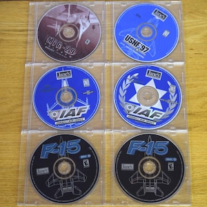 May include: A collection of six computer game CDs in clear plastic cases. The discs feature images of fighter jets and text including "MiG-29", "USNF '97", "IAF Israeli Air Force", and "F-15". The discs are in various colors including blue, black, and maroon.