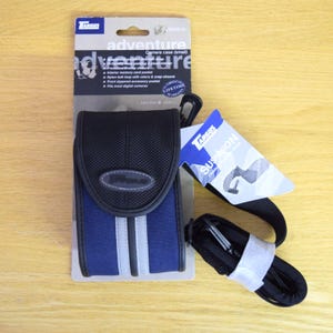 May include: A Targus small adventure camera case in blue and black with a front flap, a Targus logo patch, and two vertical grey stripes. The case is attached to a cardboard backing with product details and includes a separate black shoulder strap.