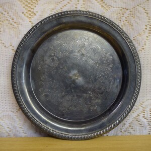 May include: A round silver-toned metal tray featuring a decorative rope-style border and an intricate, engraved scrollwork pattern centered on the flat surface. The metal has a tarnished finish with visible signs of age and wear across the plate.