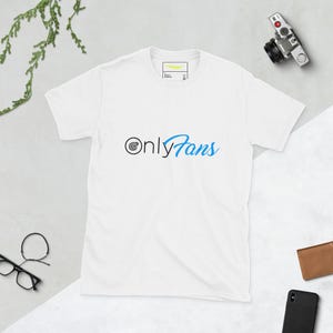 Puede incluir: Camiseta blanca con el texto "OnlyFans" en negro y azul. La camiseta tiene cuello redondo y mangas cortas. El texto está centrado en la parte delantera de la camiseta.