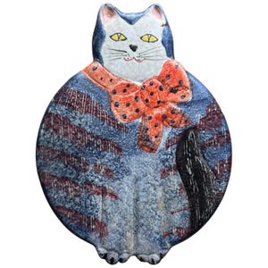May include: A ceramic cat figurine with a blue and white head, yellow eyes, and a red bow tie with black polka dots. The cat's body is blue with red and white accents, and a black tail.
