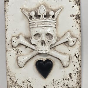 Könnte beinhalten: Rechteckige, cremefarbene Tafel mit einem gekrönten Totenkopf, gekreuzten Knochen und einem schwarzen Herz. Schädel und Knochen sind knochenweiß, mit einem gealterten, abgenutzten Aussehen. Gothic-Dekoration.