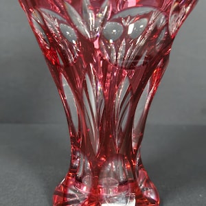 May include: A ruby red crystal vase with a flared top and a round base. The vase features intricate cut glass patterns, creating a textured and decorative surface. The vase is approximately 8 inches tall.