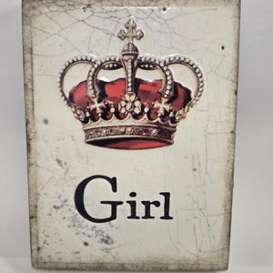 Puede incluir: Una baldosa decorativa rectangular, blanquecina, con un diseño de corona roja y dorada. La palabra "Girl" está impresa en negro debajo de la corona. La baldosa tiene un acabado envejecido y agrietado.