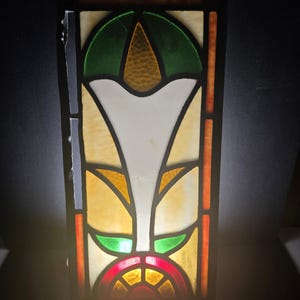 May include: A rectangular stained glass panel with a floral design. The panel features a symmetrical pattern with green, white, yellow, orange, and red glass pieces. The design includes a central white shape, green accents, and a red semi-circle at the bottom.