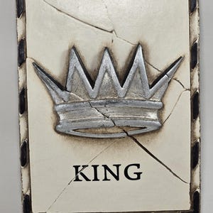 Puede incluir: Placa rectangular con un diseño de corona plateada y la palabra "KING" en negro. La placa tiene un fondo color crema con grietas y un borde oscuro con detalles negros. Decoración.