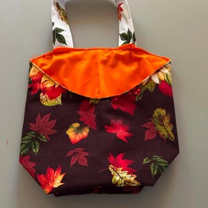 May include: A fabric tote bag featuring a dark brown background with a colourful pattern of autumn leaves in shades of red, orange, and green. The bag has a bright orange interior lining and two white handles decorated with a matching leaf print pattern.