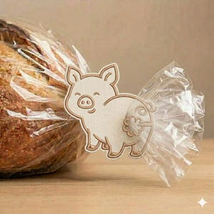 May include: A decorative pig-shaped tag with a smiling face and a floral design on its rear. The tag is attached to a clear plastic bag holding a loaf of bread. The pig has a light beige color with brown outlines.