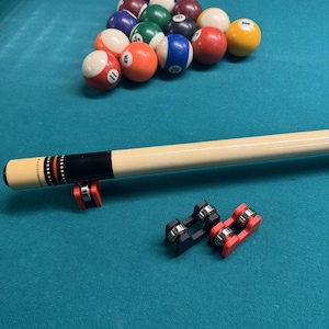 May include: A pool table with a rack of billiard balls in a triangle formation. A pool cue and two cue holders are also visible. The balls are various colors, including red, yellow, blue, and green. The table is covered in teal felt.