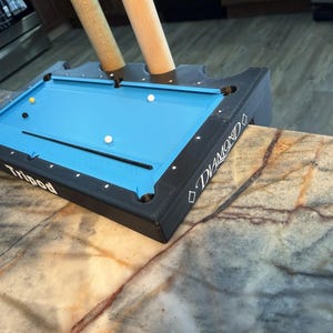 May include: A miniature pool table with a blue playing surface and black trim. The table has pockets, white and colored billiard balls, and a cue stick. The text "Tripod" is visible on the side of the table, and the word "Diamond" is also visible.