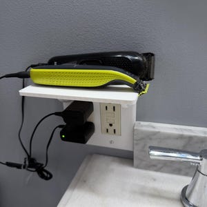 May include: A white shelf with two black and yellow electric hair clippers. The clippers are plugged into a wall outlet with two black charging adapters. The shelf is mounted on a gray wall.