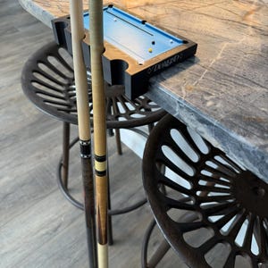 May include: A miniature pool table with a blue felt surface and a black wooden frame, along with two pool cues. The table is labeled "Diamond". The scene is set on a marble countertop with two metal stools.
