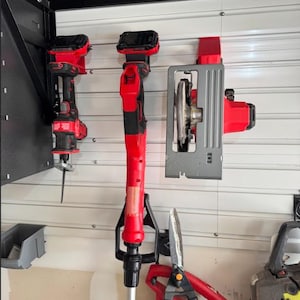 May include: A collection of red and black power tools mounted on a white horizontal slat wall. The tools include a reciprocating saw, a long-reach pole tool, and a circular saw, alongside a pair of red handled shears and a partial view of a tool.
