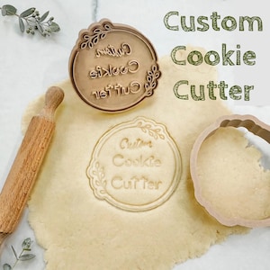 May include: A round metal cookie stamp with a floral border and the text Custom Cookie Cutter, a matching circular cookie cutter, and a wooden rolling pin resting on a sheet of rolled dough. The text Custom Cookie Cutter is also visible.