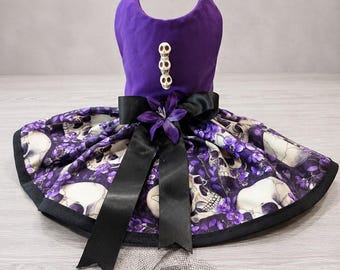 May include: A purple pet dress featuring a skull and purple floral print skirt with a black satin ribbon and a purple fabric flower at the waist. The bodice has three small skull-shaped buttons and a matching bow accessory with black netting.