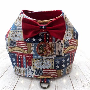 May include: A patriotic dog harness featuring a red bow tie and a decorative emblem. The harness is adorned with American flags, stars, and patriotic text. The design incorporates red, white, and blue colors, celebrating the 250th anniversary of the United States.