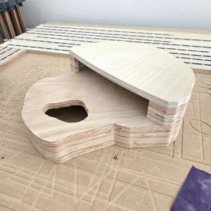 May include: A light-colored, unfinished plywood structure composed of multiple stacked layers. The object features a flat, semi-circular top platform supported by a base with a distinct, irregular cutout hole on the left side.