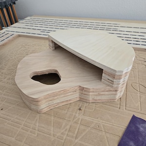 May include: A layered wooden guitar-shaped object with a cutout and a raised platform. The object is made of light-colored plywood and sits on a textured surface. A piece of purple sandpaper is visible.