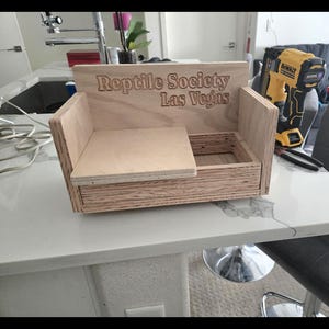 May include: A wooden reptile habitat with the text "Reptile Society Las Vegas" engraved on the front. The habitat has a rectangular base with a raised back and a sliding shelf. A yellow and black power tool is visible in the background.
